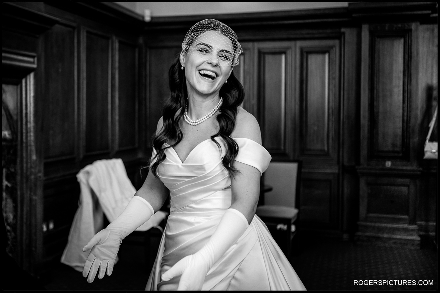 A black and white candid photograph capturing a bride's infectious laughter during her final preparations in an elegant wood-panelled suite.