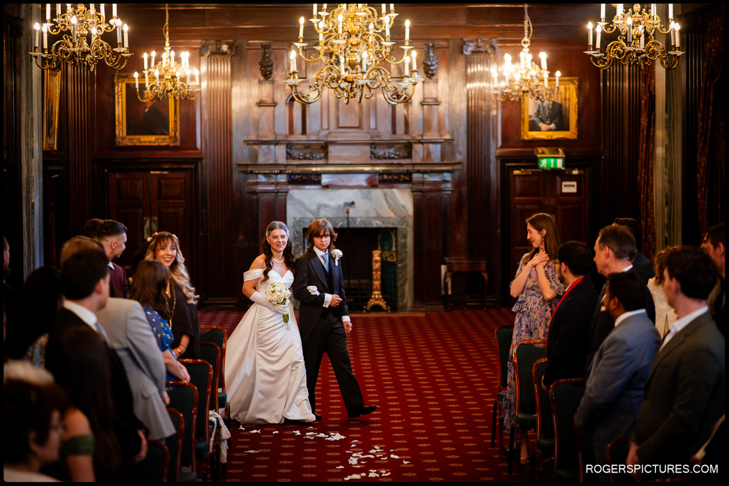 A wide documentary photograph of a bride being escorted down the aisle of a grand, historic hall filled with guests and lit by ornate chandeliers.