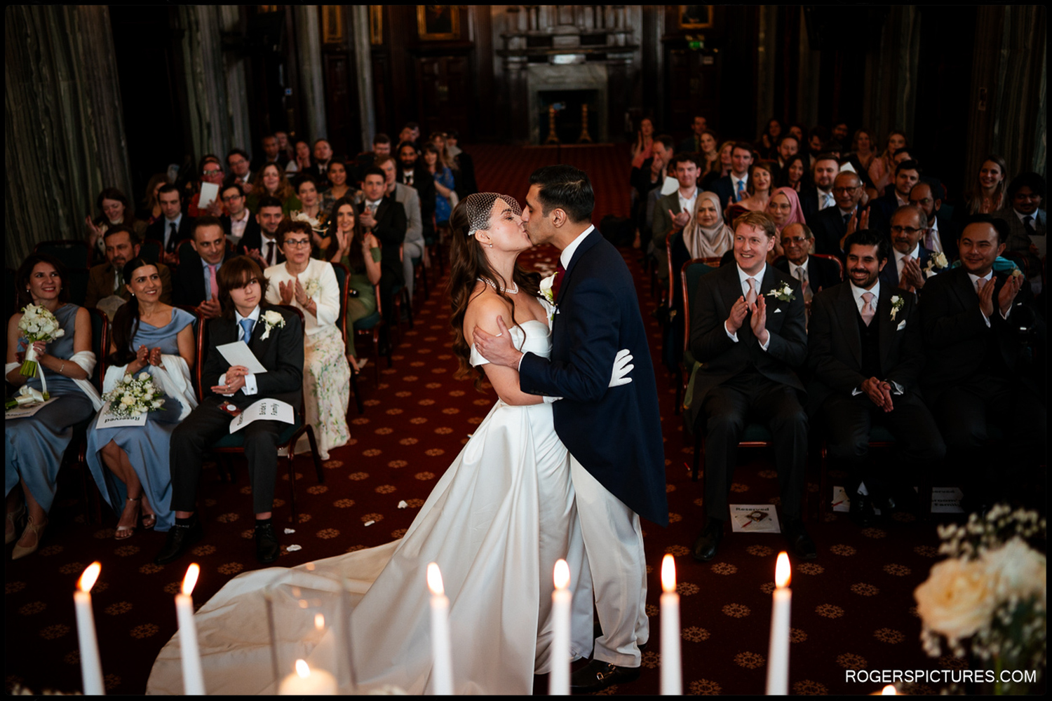 A joyful candid moment of a bride and groom sharing their first kiss at the altar, surrounded by applauding guests in the historic London venue.