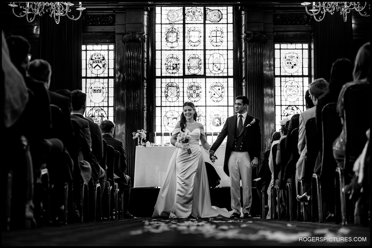 A black and white documentary photo of the newlyweds walking hand-in-hand down the aisle, framed by the silhouette of their guests and a large stained-glass window.