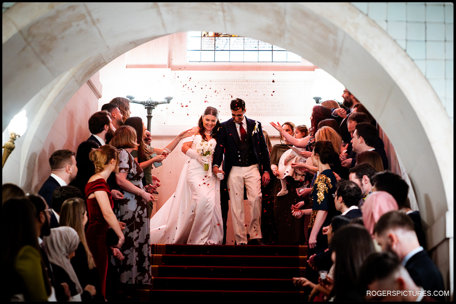 A documentary photograph captured through an archway of a couple smiling as red confetti is thrown by guests on a grand staircase.