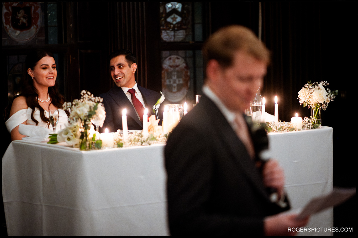 A candid shot of the newlyweds laughing at the top table during the wedding speeches at The Law Society, with a blurred guest in the foreground.
