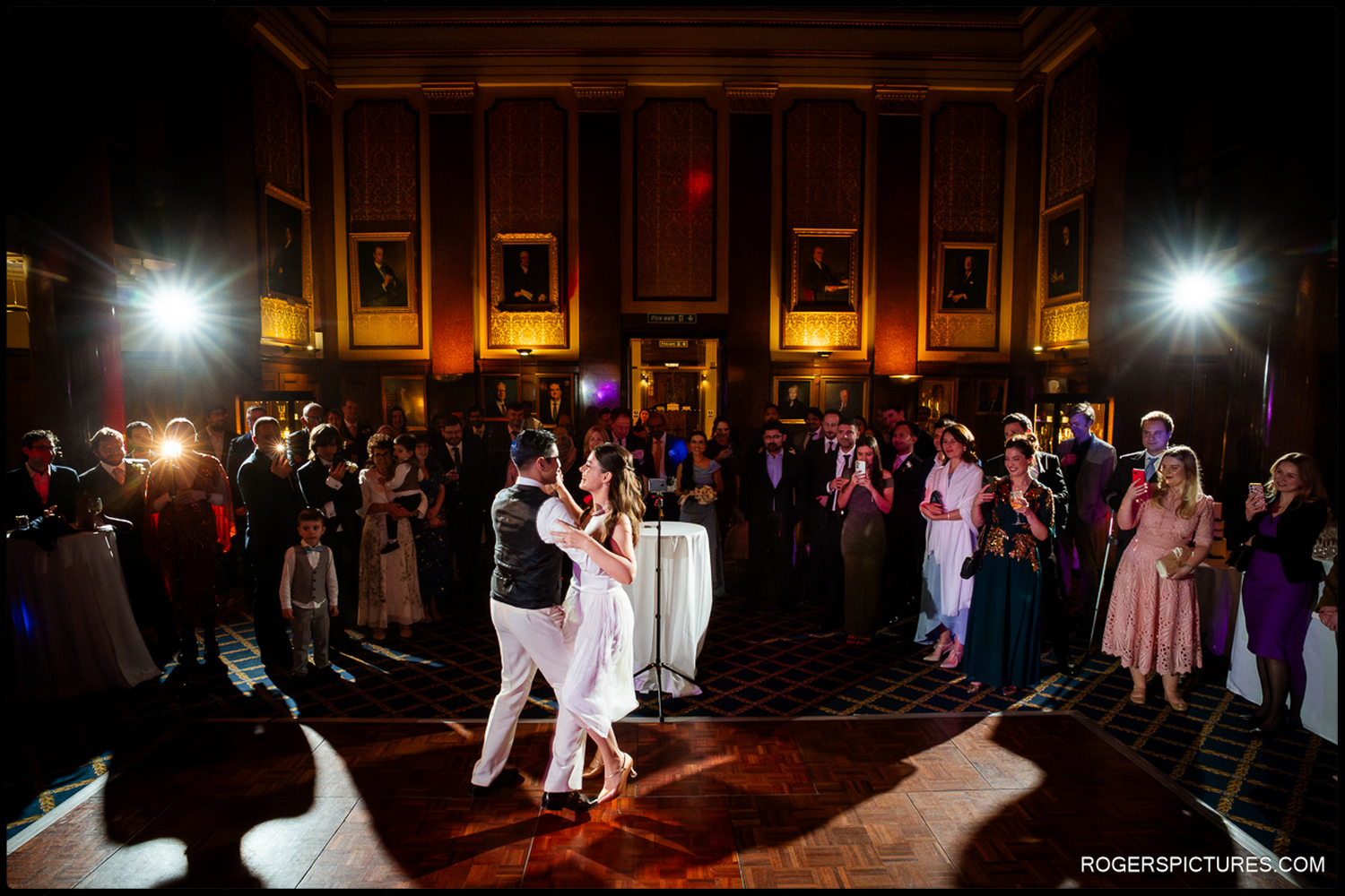 A wide-angle documentary photograph capturing the bride and groom's first dance in the grand, historic hall of The Law Society, surrounded by their guests.