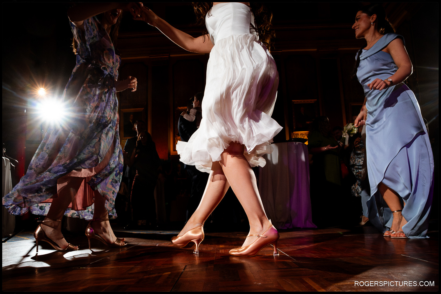 A low-angle documentary photo focusing on the movement of the bride's dress and the guests' shoes as they dance together during the wedding celebrations.